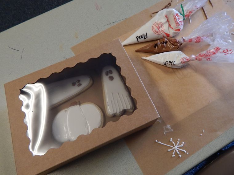 A bakery box holding ghost and pumpkin sugar cookies with piping bags beside it